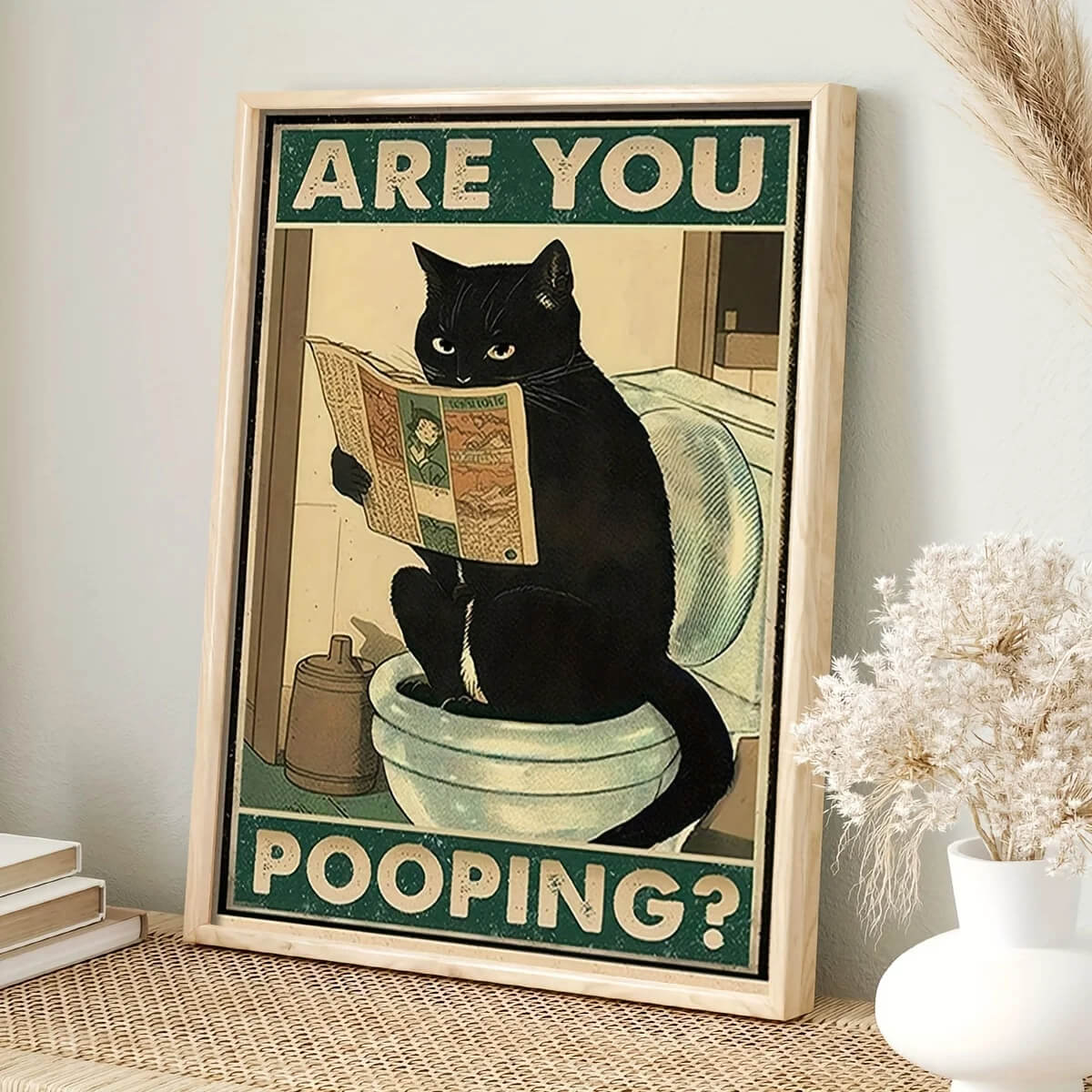 A framed canvas wall art print with a humorous depiction of a black cat sitting on a toilet, holding a newspaper, with the text 'ARE YOU POOPING?'.