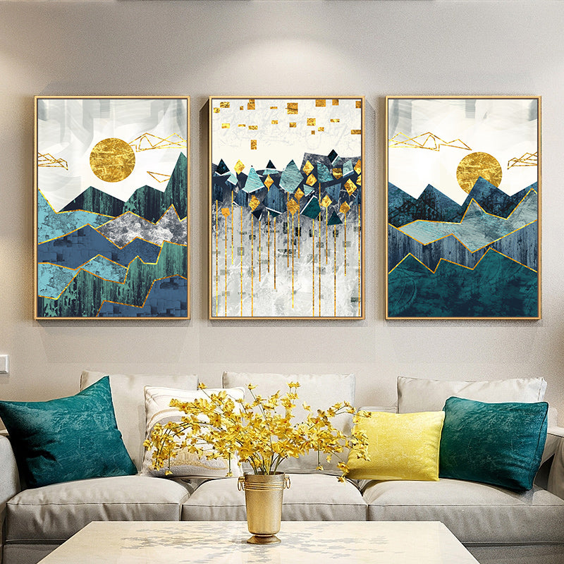 A set of three canvas prints featuring an abstract mountain landscape with blue, green, and golden colors, displayed above a grey sofa with decorative pillows.