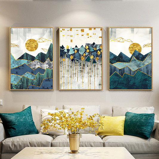 A set of three canvas prints featuring an abstract mountain landscape with blue, green, and golden colors, displayed above a grey sofa with decorative pillows.