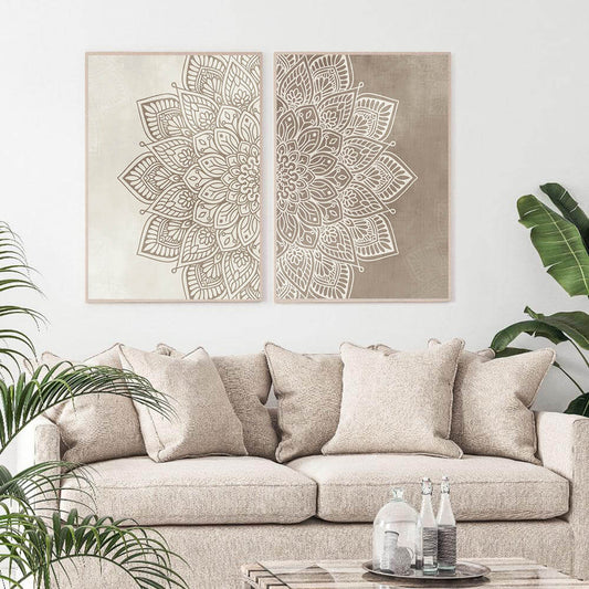 Two beige and brown mandala canvas prints hanging on a wall above a beige sofa with decorative cushions, with a plant and a glass of water on the table in the foreground.