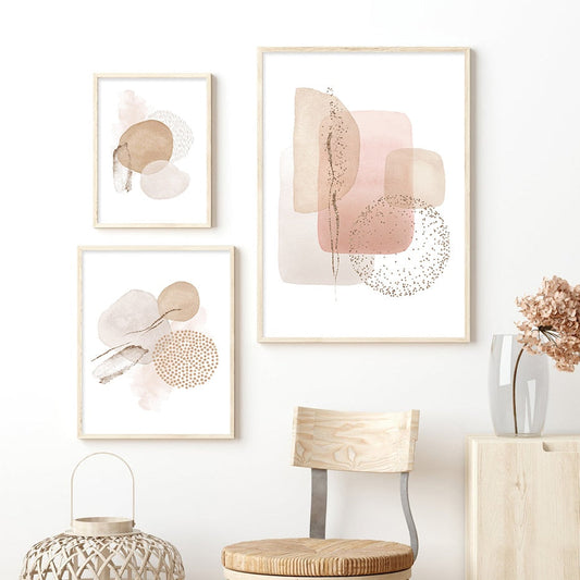 A set of three framed canvas prints featuring abstract geometric patterns in pink and beige tones, displayed above a chair and a small round table in a home setting.