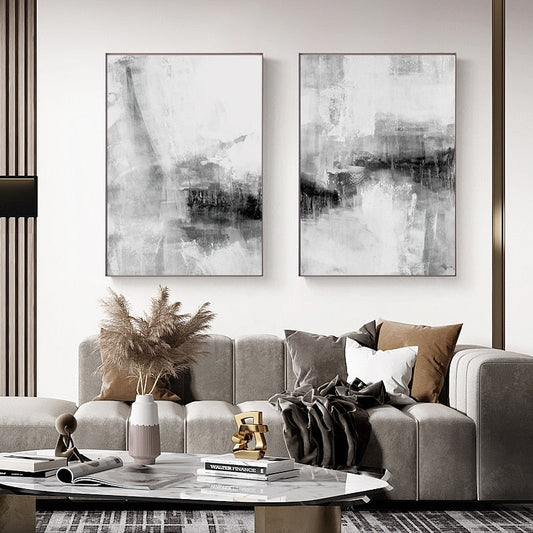 Two black and white abstract canvas prints hanging on a wall above a grey sofa with decorative pillows, a coffee table, and a decorative plant.